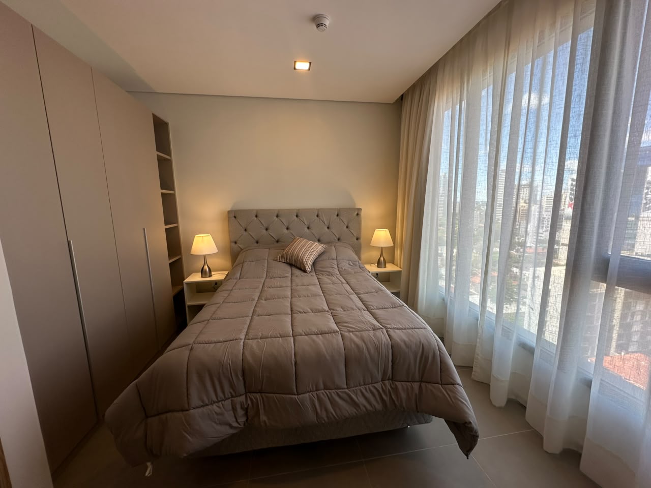 Dormitorio principal con cama tapizada, mesas de luz con lámparas y cortinas de lino frente al ventanal.
