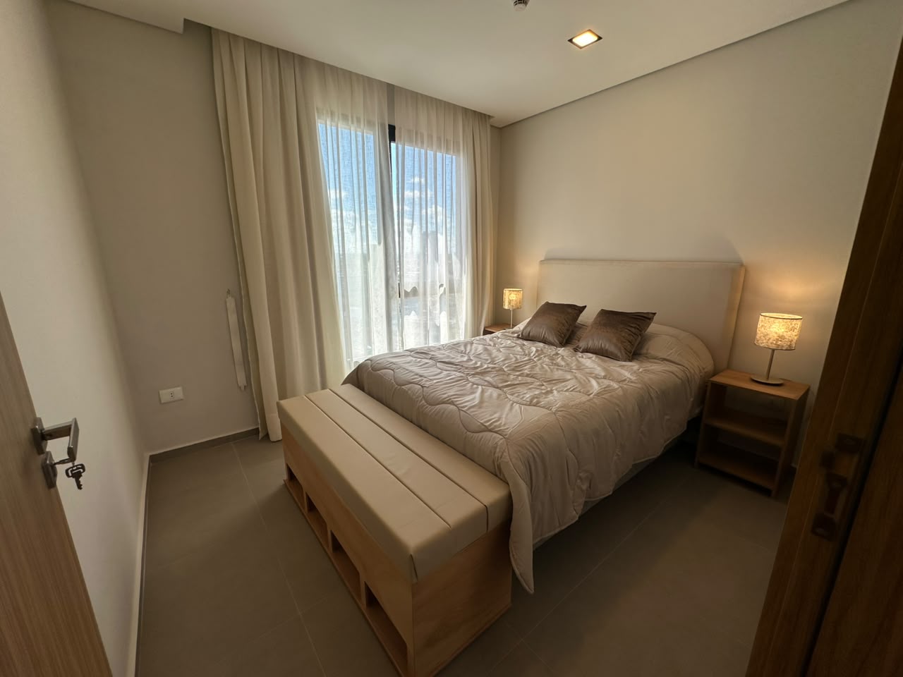 Segundo dormitorio con banco a los pies de la cama, cojines y mesa de luz con lámpara.